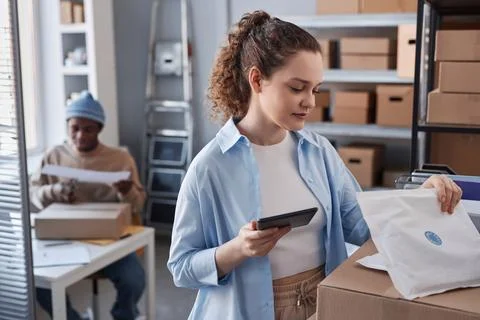 Young woman in casual attire revising codes on parcels in warehouse Stock Photos