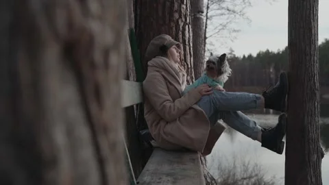 Young woman in coat is sitting on bench in pine forest with dog on her lap Stock Footage 158385853