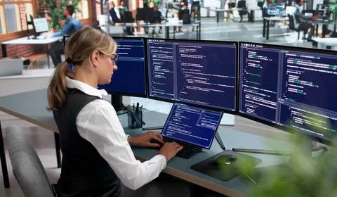 Young Woman Coding Secure Software Systems On Laptop At Busy Office Desk. Stock Photos