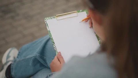 Young woman creating art while seated outdoors in city. Casual female sketcher Stock Footage 346543577