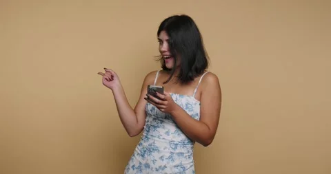 Young woman dances while looking at her phone in a studio with a plain back.. 스톡 동영상 326793065