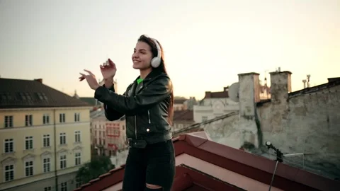 Young woman dancing on a rooftop at sunset, enjoying music through wireless Stock Footage 320906265