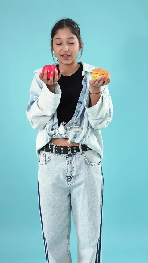 Young Woman Deciding Between Apple and Hamburger on Blue Background Vídeo Stock 345538721