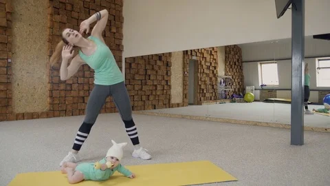 Young woman does leaning exercises and squats next small child indoors Stock Footage