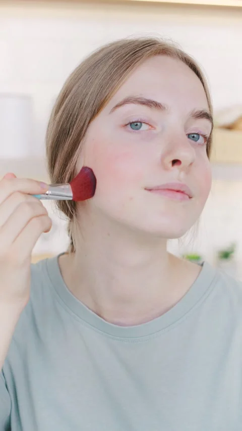 Young woman doing makeup applying blush with a brush in a bright room during the Stock Footage 318556431