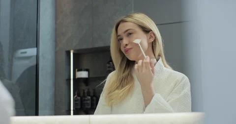 Young woman doing makeup using brush in front of mirror in bathroom. Enjoying Stock Footage 273088761