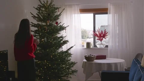 Young woman drinking coffee and looking at decorated Christmas tree in cozy room Stock Footage 324924012