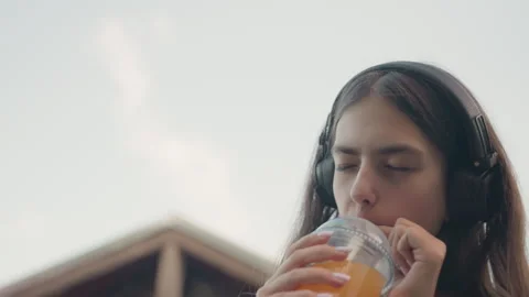 Young Woman Drinking Juice While Wearing Headphones In Gym With Outdoor Elements Stock Footage 345974147