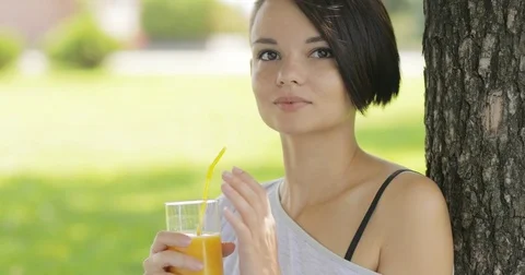 Young woman drinking orange juice near tree in summer Stock Footage