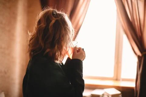 Young woman drinking tea while looking through window standing at home Stock Photos