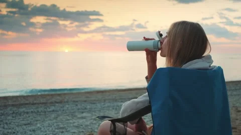 Young woman drinking from thermos while sitting on beach camping chair Video stock 342329178