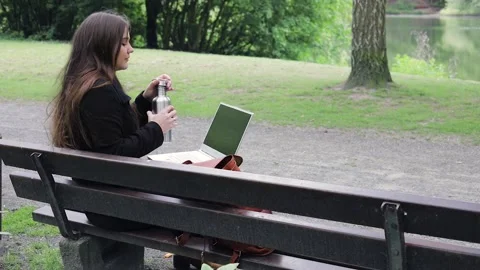 Young woman drinks water while working with laptop in a park on a bench Video stock 130397641