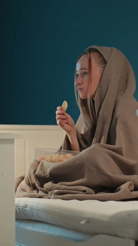 Young woman eating snacks while covered with blanket on bed in bedroom showing Stock Footage 344429327