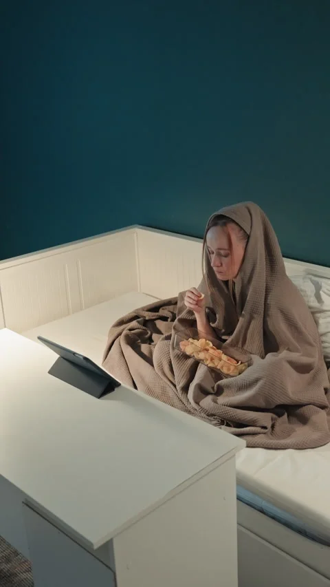 Young woman eating snacks while looking at screen under dark blue bedroom Stock Footage 344429541