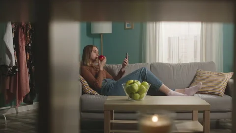Young woman eats a red apple, lying on sofa and and watching something gadget Stock Footage 149146557
