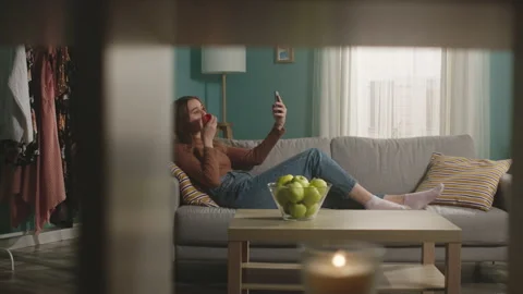 Young woman eats a red apple, lying on sofa and and taking a selfie by phone Stock Footage 151591573