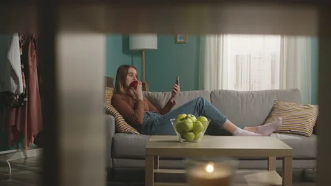 Young woman eats a red apple, lying on sofa and and speaks via video link Stock Footage 151899338