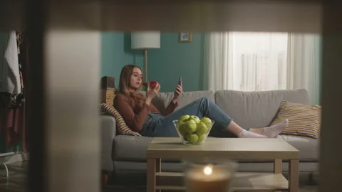 Young woman eats a red apple, lying on sofa and and speaks via video link Stock Footage 151899414