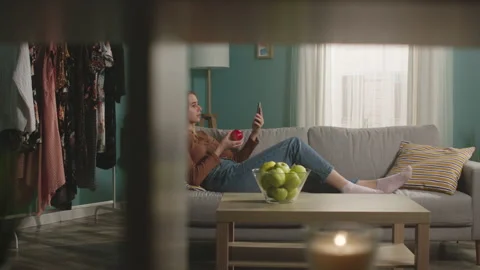 Young woman eats a red apple, lying on sofa and and speaks via video link Stock Footage 151923923