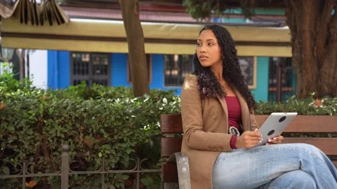 A young woman is engaged in using a tablet while sitting outdoors in an urban Vidéo 313818893