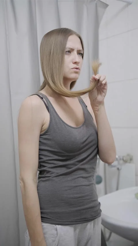Young woman examining split ends of her hair with frustration in bathroom mirror Stock Footage 312466602