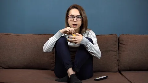 A young woman in glasses eats chips and watches TV. Girl sitting with legs on Stock Footage 118084013