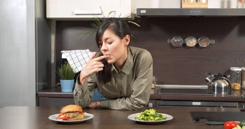 Young woman having difficulty choosing between green salad and burger in kitchen Stock Footage 149052372