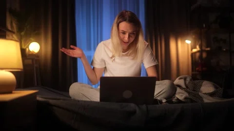 A young woman having a video call at night while sitting on her bed in a cozy Stock Footage 304086653
