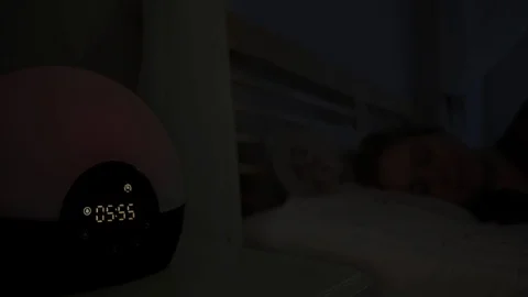 Young woman in her bed while her alarm goes off simulating sunrise Video stock 114430520