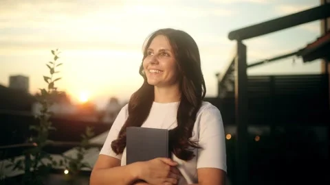 A Young Woman Holding a Book While Enjoying a Beautiful Sunset on a Rooftop Stock Footage 283576895