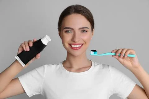 Young woman holding brush and tube with charcoal toothpaste on grey backgroun Stock Photos