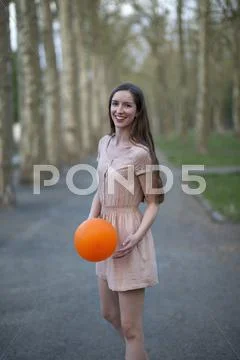 A Young Woman Holding An Orange Ball, Standing In A Park