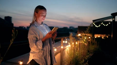 A Young Woman Joyfully Using Her Phone on a Rooftop Surrounded by String Lights Stock Footage 280987182