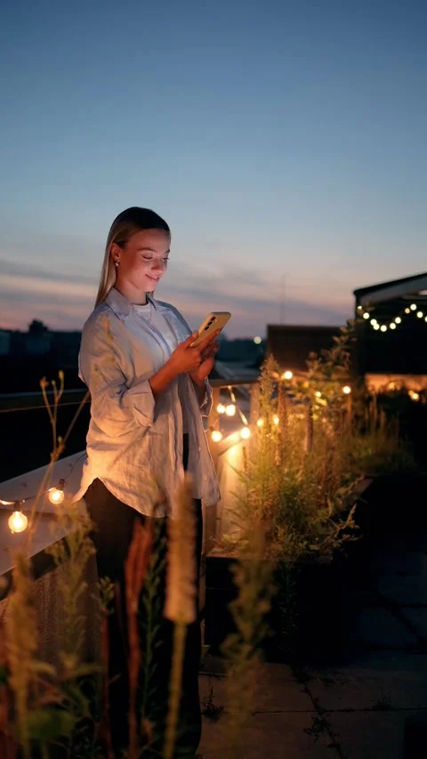A Young Woman Joyfully Using Her Phone on a Rooftop Surrounded by String Lights Stock Footage 282024394
