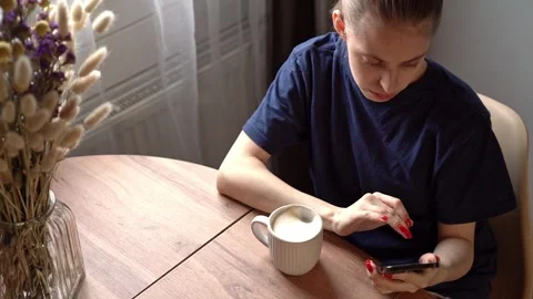 Young woman at kitchen table lowering mug beside smartphone in quiet home Stock Footage 341554629