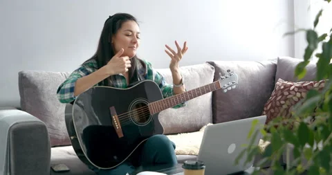 Young woman learning to play guitar online talking with teacher using laptop at Stock Footage 150746853