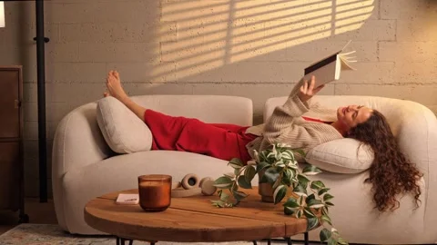 Young woman lying back on a couch reading a book in a cozy sunlit living room Stock Footage 341336171