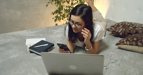 Young woman lying on the bed using a smartphone and laptop while working at home Stock Footage 127181408