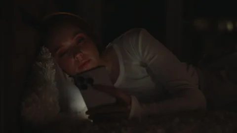 Young woman lying in bed, using smartphone at night, illuminated by soft glow of Stock Footage 302564907