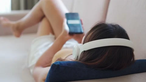 Young woman lying on sofa in living room and listening music form smart phone Видео 145384853