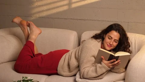 Young woman lying on sofa reading a book and turning page in cozy living room 库存影片 341336007