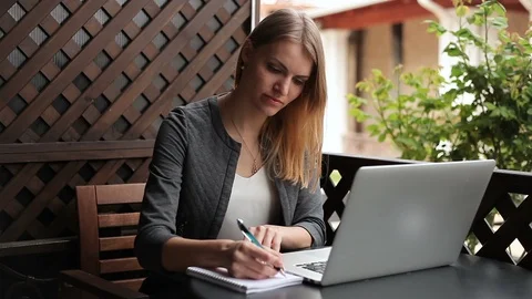 A young woman Makes notes in a notebook, working on a laptop at a cafe table on  Vidéo 114087148