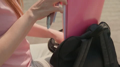 Young woman placing pink folder into black backpack outdoors during daylight Stock Footage 345975686
