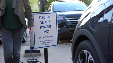 Young woman plugs connecting electric vehicle EV at dedicated parking Stock Footage