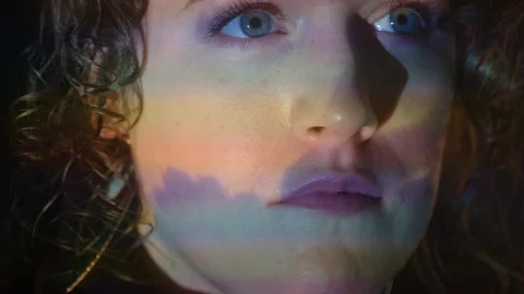 Young woman with rainbow pattern and clouds on face seems thoughtful - projector Stock Footage 101980175