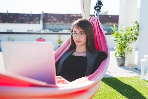 Young woman reclining in city roof terrace hammock typing on laptop Stock Photos