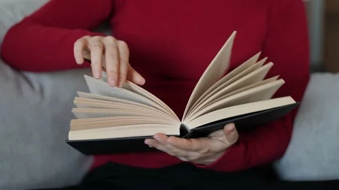 Young woman with red pullover reading hardcover book on grey couch at home. Stock Footage 142842296