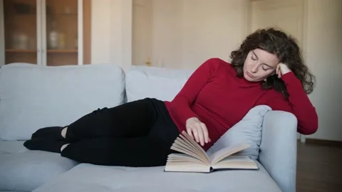 Young woman with red pullover reading hardcover book on grey couch at home. Stock Footage 142842686