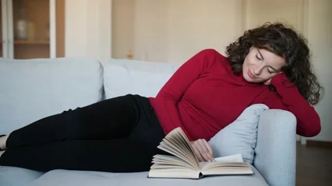 Young woman with red pullover reading hardcover book on grey couch at home. Stock Footage 142842907
