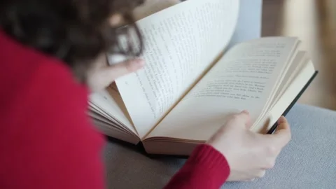 Young woman with red pullover reading hardcover book on grey couch at home. Stock Footage 142843007